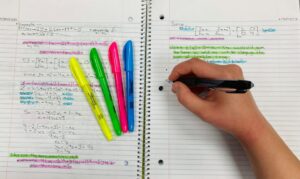 Learner writing Hebrew verb forms in a notebook beside a grammar chart and open Hebrew text.