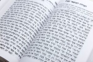 A learner reads Hebrew letters aloud beside a notebook, pencil, and open language workbook at a home desk.