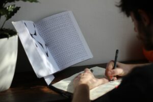 A learner writes Hebrew words in a notebook beside flashcards and an open Hebrew reading book.