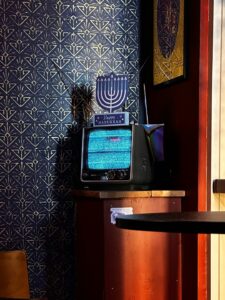 Adults watch a Hebrew comedy program with notebooks and subtitles visible on a living-room screen.