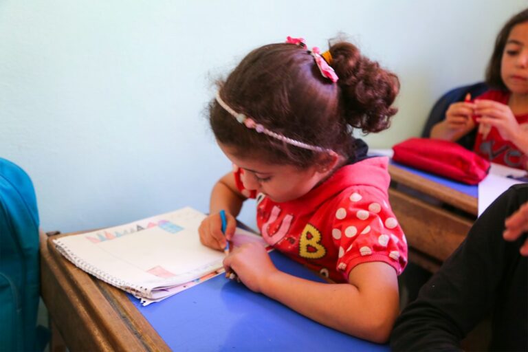 Adult student practices reading Hebrew letters from a workbook at a kitchen table.