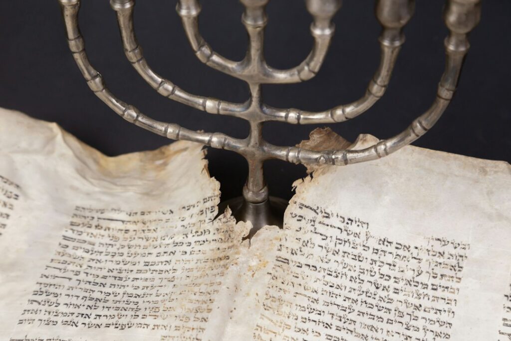 Adults and children study Hebrew letters together around a table with books, notebooks, and handwritten practice sheets.