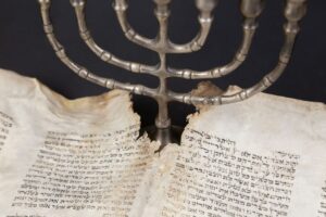 Adults and children study Hebrew letters together around a table with books, notebooks, and handwritten practice sheets.