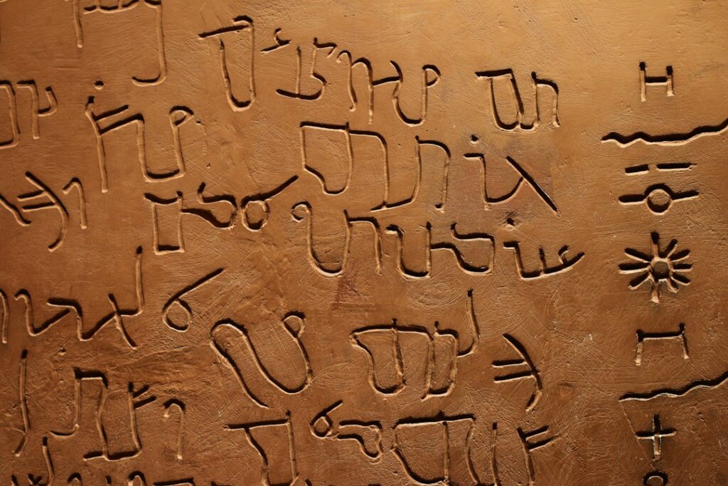Person writing a personal name in Hebrew letters beside an open Hebrew alphabet guide.