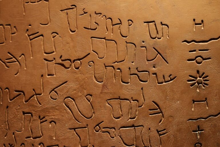 Person writing a personal name in Hebrew letters beside an open Hebrew alphabet guide.